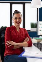 Portrait of entrepreneur sitting at desk in startup office. Businesswoman in red shirt looking confident in business workspace. Successful employee posing with crossed arms in front of desktop computer. photo
