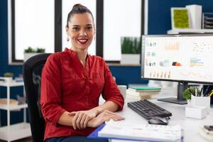 Portrait of smiling small business owner in start up office. Entrepreneur in red shirt looking confident at desk with clipboard and desktop computer. Manager posing with turnover results. photo