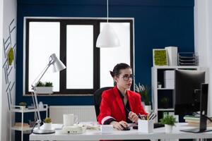 Businesswoman at desk in startup office enviroment. Small business owner with glasses working at desktop computer. Employee in red jacket sitting in blue and white workspace with big window. photo