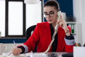 Entrepreneur taking notes in phonecall sitting at desk. Businesswoman with glasses working in startup office. Employee in red jacket writing on paper in telephone conversation. photo
