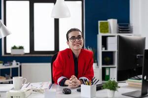 Portrait of businesswoman with glasses in startup office. Professional entrepreneur in red jacket looking confident in business workspace. Confident employee posing in front of desktop computer. photo