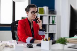 Confident small business owner talkin on smartphone sitting at desk. Employee with glasses looking at computer screen while talking on the phone. Manager in red jacket in mobile phone conversation. photo