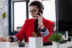 Businesswoman taking notes in smartphone phonecall sitting at desk. Entrepreneur with glasses working in startup office. Small business owner in red jacket writing on paper in startup enviroment. photo