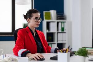 Businesswoman with glasses working at desk in startup office. Confident manager in red jacket typing on computer keyboard. Focused start up owner at workplace using desktop pc. photo