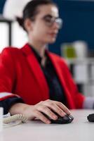 Closeup of hand using wireless computer mouse in startup office. Businesswoman with glasses scrolling content on desktop computer. Entrepreneur in red jacket clicking in business workspace. photo