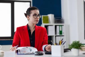 Businesswoman at startup office using clipboard with chart. Entrepreneur with glasses working at business desk. Focused employee in red jacket holding pen looking at computer screen. photo