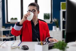 Employee with glasses at startup desk drinking from white cup. Businesswoman in red jacket having coffee or tea break. Small business owner taking a pause from working with computer. photo