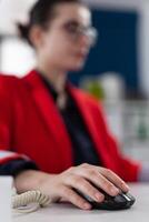 Closeup of businesswoman using wireless computer mouse in startup office. Entrepreneur with glasses clicking in business workspace. Entrepreneur in red jacket scrolling content on desktop computer. photo