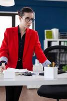 Small business owner standing up next to desk holding pen and papers. Focused businesswoman with glasses and red jacket in startup office looking at business results on computer screen. photo