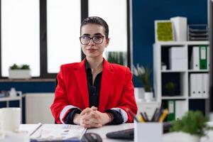Portrait of entrepreneur with glasses in startup office. Professional businesswoman in red jacket looking confident in business workspace. Confident employee posing sitting at desk. photo