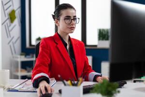 Small business owner with glasses working at desk in startup office. Businesswoman with glasses working in startup worpklace. Focused entrepreneur in red jacket looking at computer screen. photo