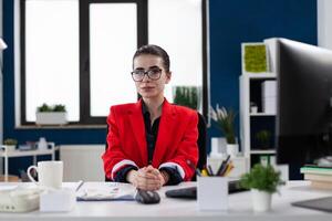 Portrait of small business owner with glasses in startup office. Professional employee in red jacket looking confident in business workspace. Confident businesswoman posing sitting at desk. photo