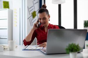 Successful manager in red shirt talking on smartphone. Smiling entrepreneur explaining charts on the mobile phone at startup desk with laptop computer. Businesswoman happy with business results. photo