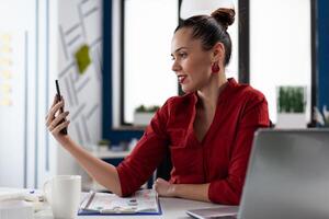 Entrepreneur in casual videocall conference using smartphone. Smiling businesswoman in red shirt using mobile phone in startup business office. Startup manager looking at content on social media. photo