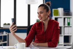 Smiling businesswoman in red shirt looking at content on social media. Entrepreneur in casual videocall conference using smartphone. Startup manager using mobile phone in startup business office. photo