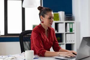 Confident businesswoman in red shirt working at desk in startup office. Business manager typing on laptop keyboard. Focused start up owner at workplace using notebook computer. photo