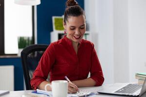 Entrepreneur writing notes on clipboard sitting at desk. Businesswoman with red shirt working in startup office in front of laptop. Smiling employee analyzing startup business chart. photo