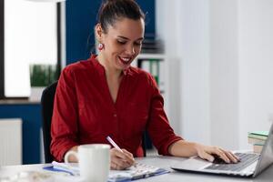 Businesswoman writing notes on clipboard with one hand on laptop. Smiling entrepreneur with red shirt sitting at desk. Employee working analyzing startup business chart while typing. photo