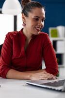 Smiling entrepreneur in red shirt looking at laptop screen. Businesswoman in call conference touching notebook computer touchpad. Small business owner watching content on social media. photo