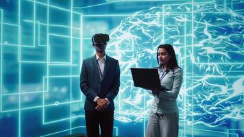 Technician wearing VR headset looks at hologram of artificial intelligence human brain simulation, assisted by colleague using laptop. Team of programmers interacting with AI visualization, camera B video