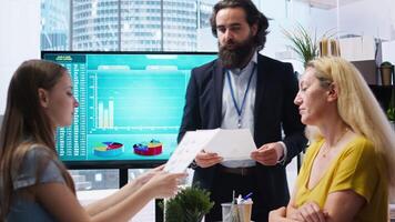 Financial advisor showing statistical graphs on big screen to customers, analyzing market trends to optimize income. Family members checking investment plan on display made by consultant, camera A video