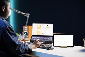 Black woman sitting at desk, tablet showing isolated white screen while she browses on her laptop. African american freelancer using her personal computer as a digital device displays mockup template. photo