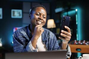 Close-up of smiling black woman grasping and looking at her mobile device. Portrait of african american female influencer using smartphone to browse the internet and surf social media. photo
