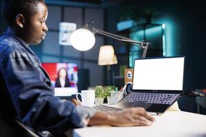 Female freelancer focused and utilizing technology while working at home with digital laptop having isolated white screen. Black woman looking at personal computer displaying blank mockup template. photo
