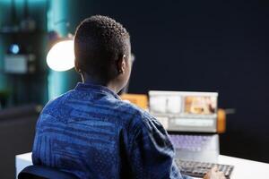 An African American woman edits footage on her laptop at her home office desk, immersed in creative work. Modern studio setup with digital device and software for productive filmmaking. photo