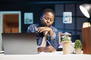 Female blogger with opened laptop, reviewing notes, and writing ideas on notepad. Black woman working from home, taking notes and utilizing technology for productivity and innovation. photo
