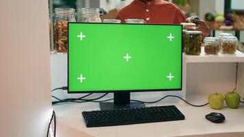 Seller at register uses greenscreen on computer, showing isolated copyspace display on pc monitor. Young man small business owner encourages healthy lifestyle with blank chromakey. photo