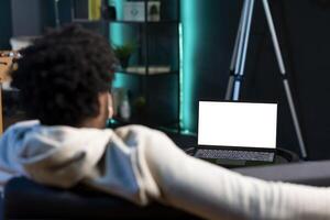 Man relaxing in apartment by watching TV series on mockup laptop. BIPOC person enjoying leisure time in neon illuminated home, looking at television shows on isolated screen notebook photo