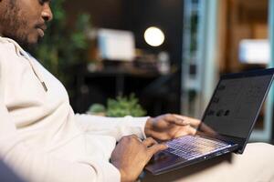 Remote worker at home on couch looking at financial graphs on laptop screen, close up shot. Man in modern stylish room trying to complete job tasks before deadline, analyzing business charts photo