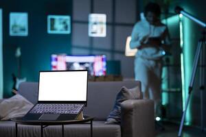 Focus on isolated screen laptop on coffee table in neon lit home with man in blurry background. Close up shot of mockup notebook and person in the back reading ebook on tablet photo