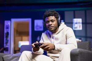Smiling BIPOC man playing shooter videogame in neon lit apartment, holding controller. Happy gamer participating in internet multiplayer game using console system and headphones photo