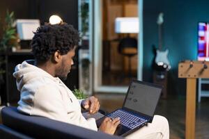 Remote worker at home on couch looking at financial graphs on laptop screen. African american man in apartment trying to complete job tasks before deadline, analyzing business charts photo
