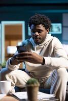 Man texting internet friends using mobile phone in warm stylish home, having fun. Joyful person relaxing in apartment by scrolling on smartphone, entranced by social media activity photo