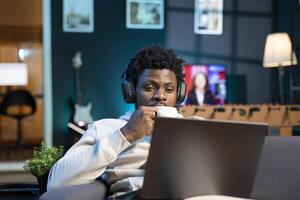 Self employed man drinking coffee and writing emails at home while listening music. African american remote employee doing b2b networking over internet, wearing headphones, enjoying hot beverage photo