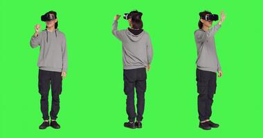 Young adult enjoys virtual reality gadget in studio with greenscreen background, using interactive vision or simulation on headset. Modern guy having fun with artificial intelligence vr glasses. photo