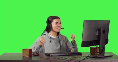 Asian guy sitting at office desktop, having fun with friends while enjoying internet multiplayer games with headphones. Over full body greenscreen, man competes in rpg contest. photo