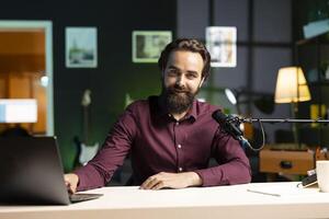 Portrait of smiling man in studio looking at videos on laptop sent by viewers, filming with professional camera. Cheerful entertainer reacting to clips sent by audience during live broadcast photo