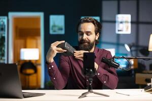 Tech expert presenting features of newly launched V mount battery, filming technology review using smartphone on tripod. Reviewer showcasing charging device specs, recording with phone photo