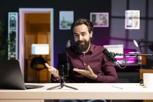 Smiling media star using smartphone on tripod to film vlog channel intro for online streaming platforms. Cheerful man greets viewers, presenting today topic, recording with phone photo