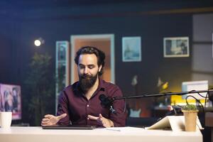 Man in studio endorsing laptop from brand sponsoring , talking in professional microphone. Content creator does influencer marketing by promoting notebook, recording with high tech mic photo