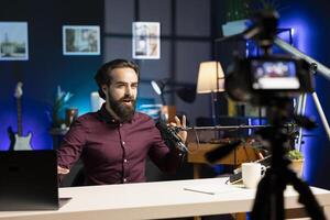 Influencer talking in professional microphone in studio illuminated with rgb lights at night, filming internet production. Man uses content creation recording mic in apartment photo