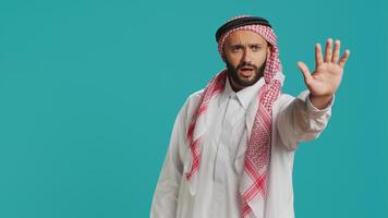 Middle eastern guy doing stop gesture with palm, expressing negativity and refusal in studio. Young male model presenting rejection, being disappointed with stuff over backdrop. photo