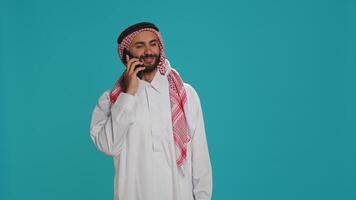 Islamic guy in robe texting on phone while posing with confidence over blue background. Middle eastern person dressed in thobe and headscarf, sending online messages on smartphone app. photo