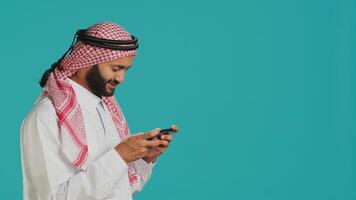 Arabic person playing mobile videogames on smartphone with tensed expression, wears islamic headscarf and thobe. Muslim gamer enjoying online gaming experience on phone app in studio. photo