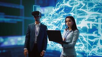 Technician wearing VR headset looks at hologram of artificial intelligence human brain simulation, assisted by colleague using laptop. Team of programmers interacting with AI visualization, camera B photo