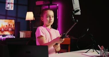 Child filming with smartphone attached to selfie stick in living room used as professional studio for recording vlogs. Happy kid captures footage with phone in neon lit apartment, discussing topics photo